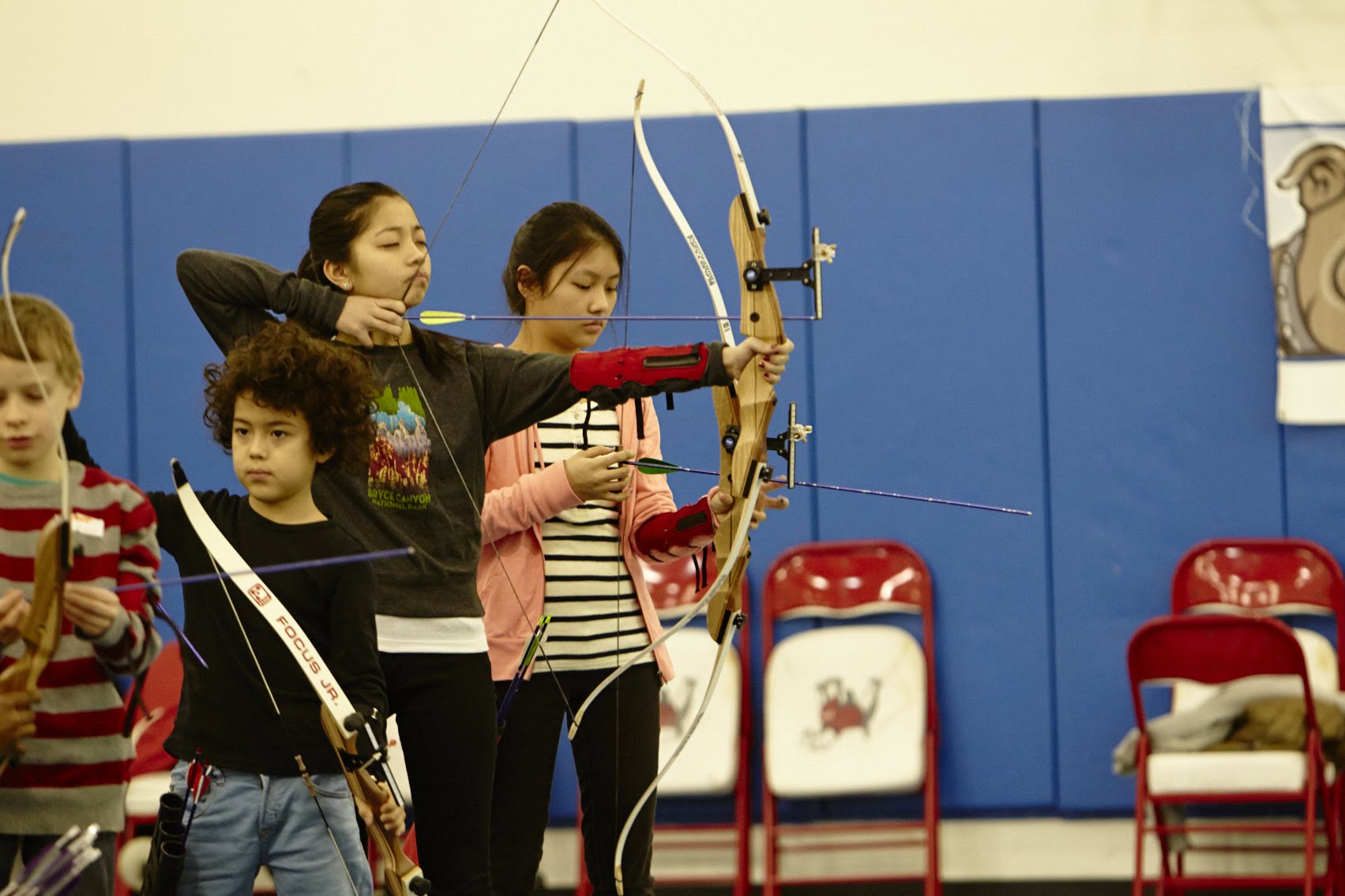 Kids practicing archery with instructors in a school gym